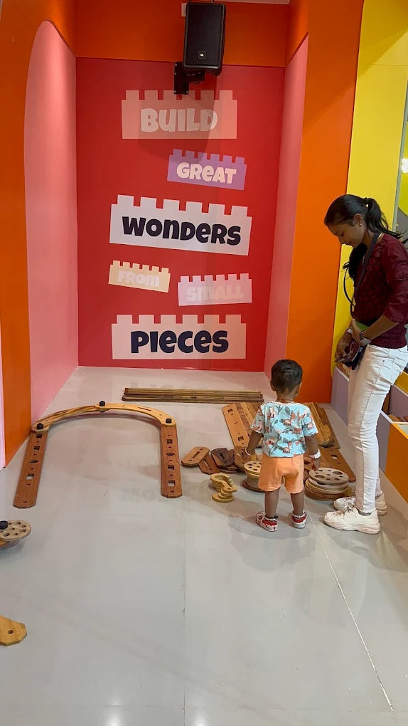 A young child and an adult stand in a brightly colored room with building blocks and wooden toys. The wall behind them has large text that says "BUILD GREAT WONDERS FROM SMALL PIECES."