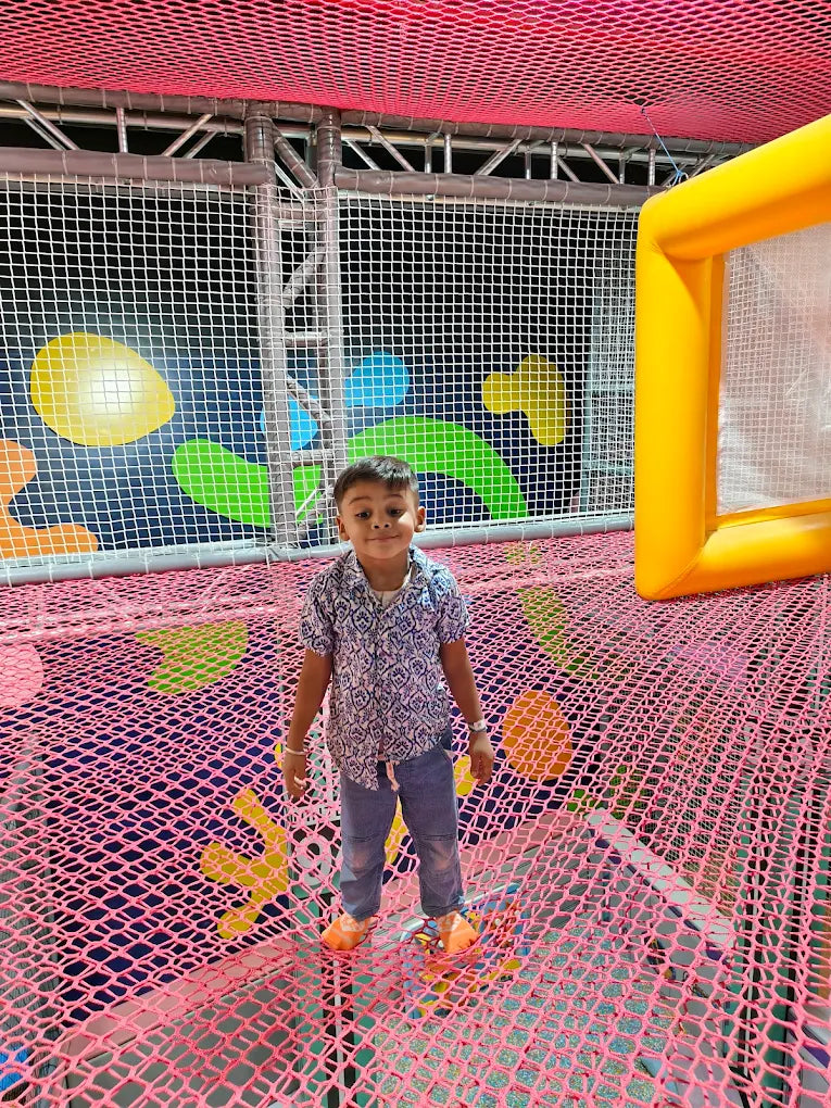 A young boy stands on a pink net floor in a colorful indoor playground.