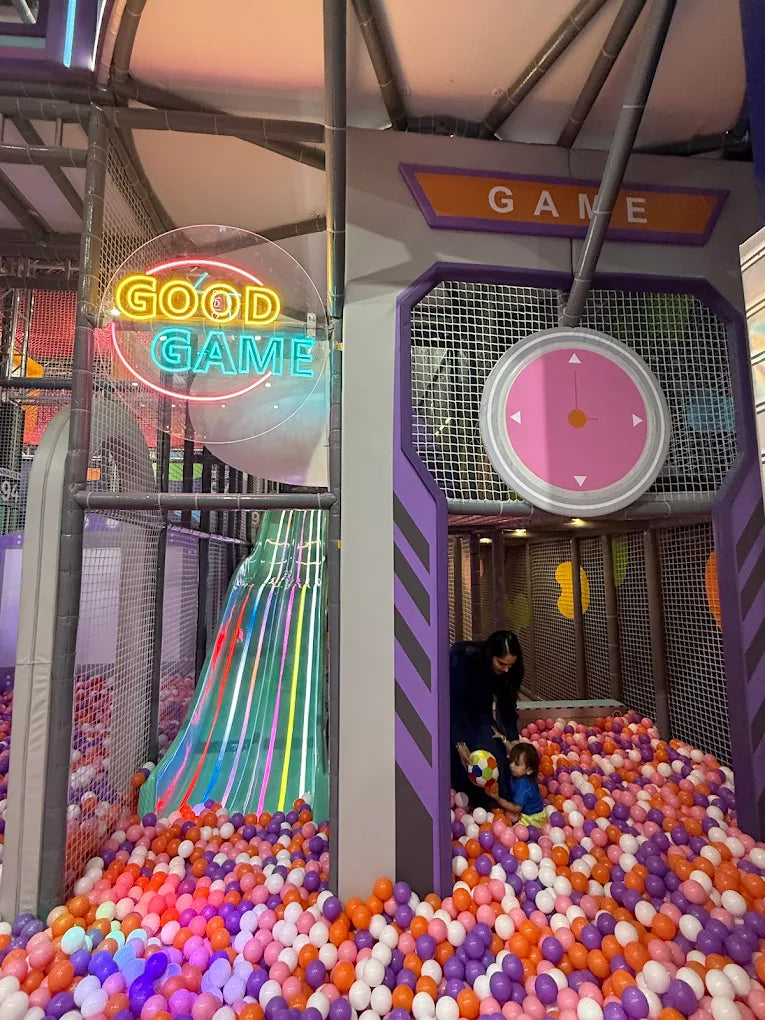 A woman and a young child play in a ball pit with a colorful slide. Neon signs read "GOOD GAME" and "GAME".