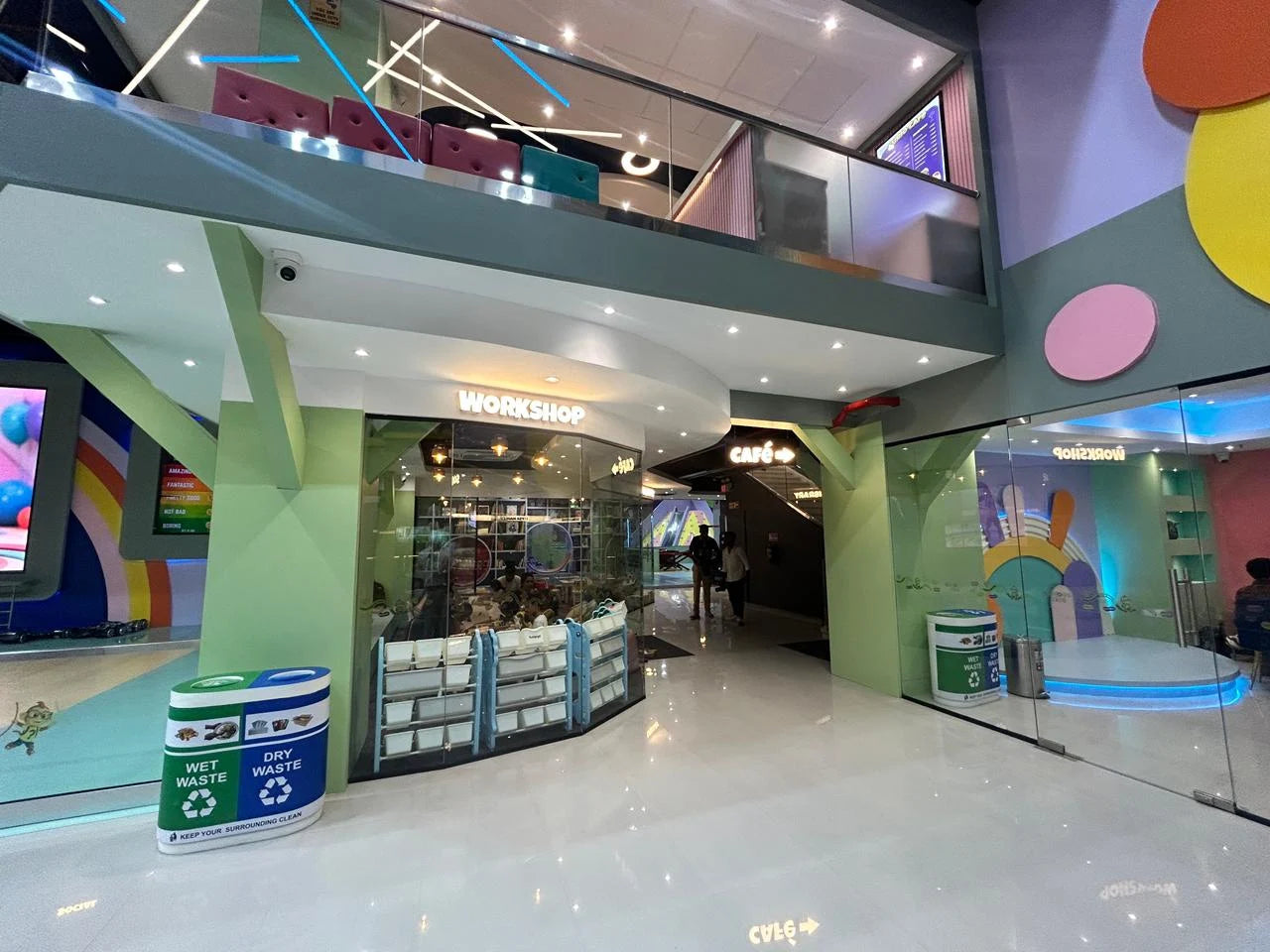 Interior of a modern, colorful indoor play space with a "Workshop" and "Cafe" area, and children playing.