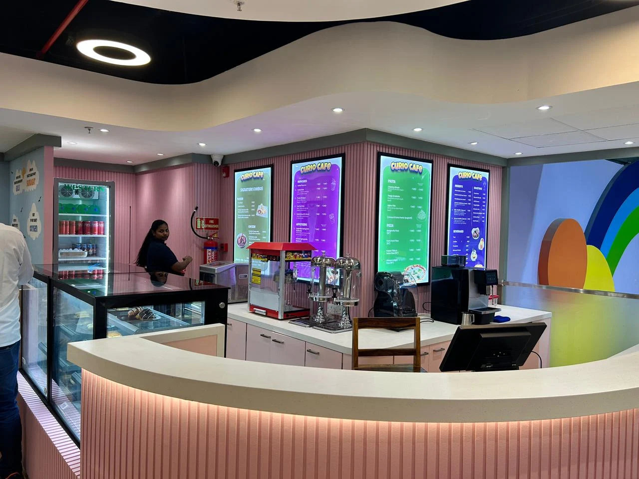 A young woman stands behind a counter in a brightly lit cafe with colorful menu boards.