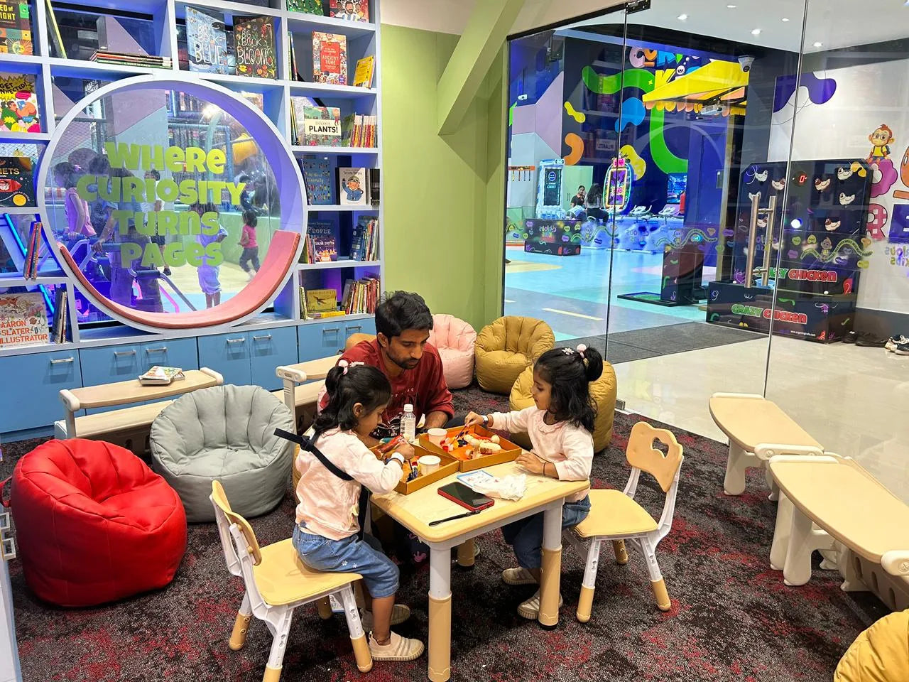 A father and two young daughters play at a table in a colorful, child-friendly library.