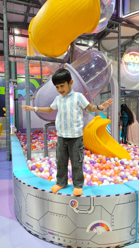 A young boy balances on the edge of a ball pit in a colorful indoor playground. He is wearing a striped shirt and cargo pants.