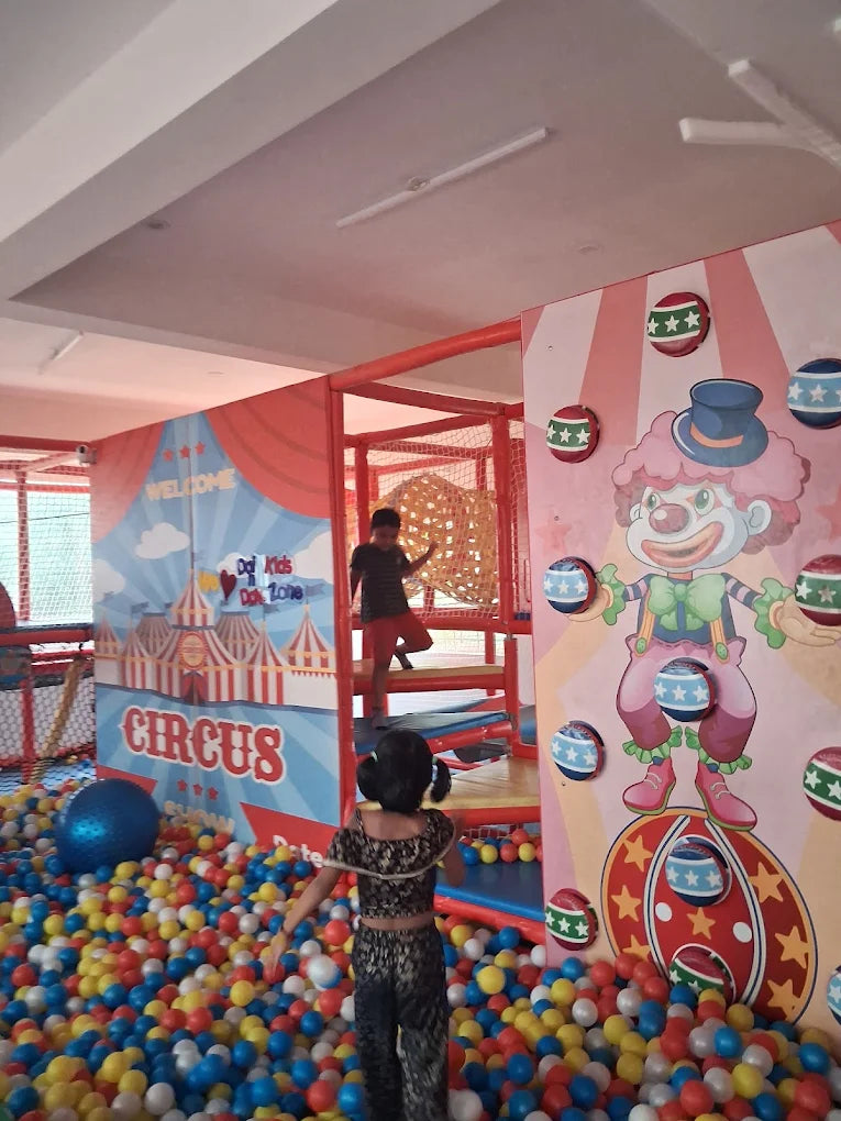 Children play in a colorful indoor circus-themed play area with a ball pit and climbing structures.