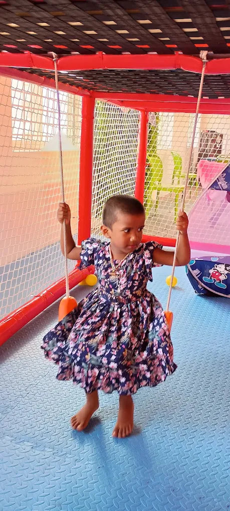 A young child in a floral dress swings on a swing set in a colorful indoor play area.