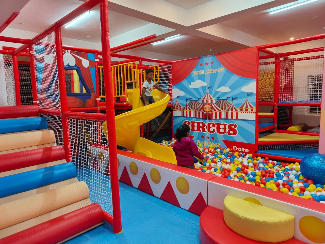 Children play in a colorful indoor circus-themed playground with a ball pit and slide.