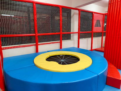 A colorful indoor playground with a trampoline surrounded by blue padding and red netting.