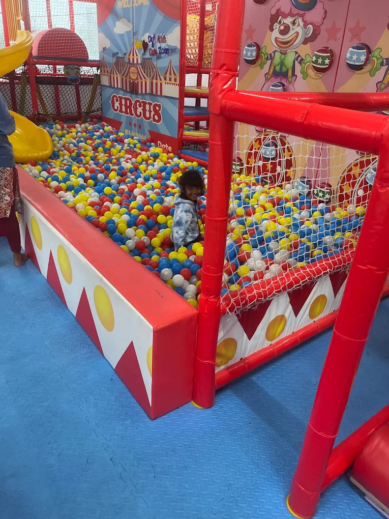 A young child plays in a ball pit at a circus-themed indoor playground.