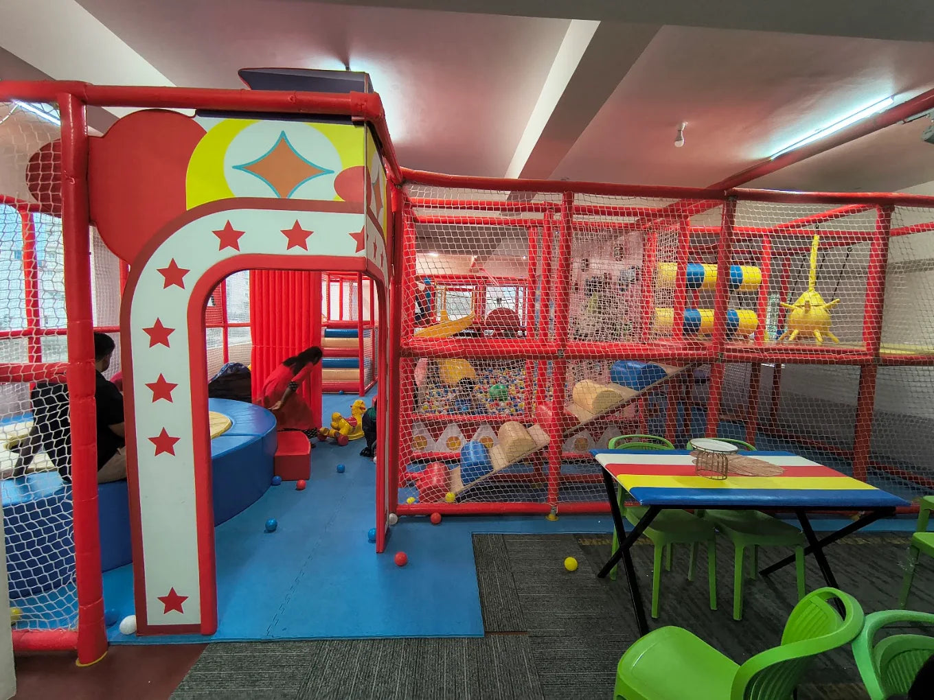 A colorful indoor playground with children playing in a ball pit and a table with chairs.