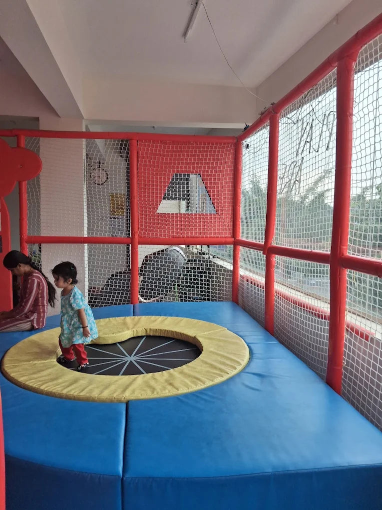 A young child jumps on a trampoline in a soft play area while an adult watches.