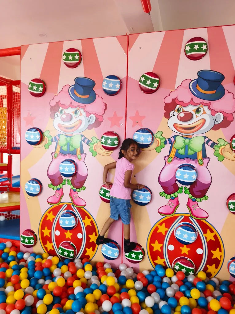 A young girl plays in a ball pit in front of a clown-themed wall with colorful balls.