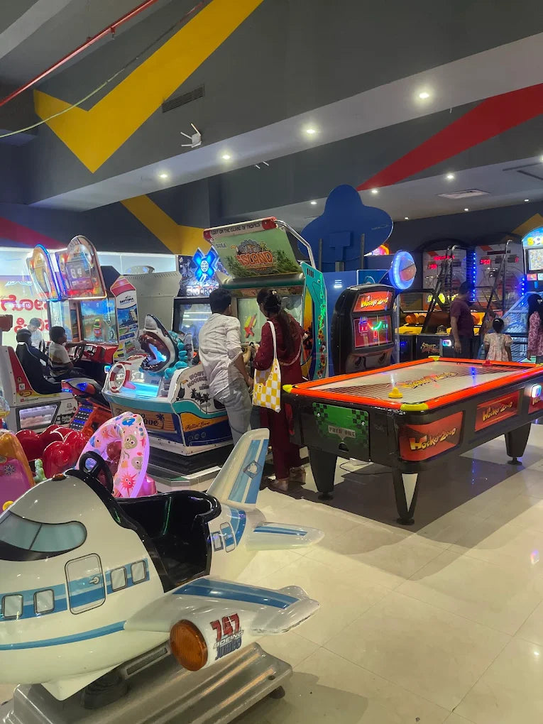 People play arcade games and air hockey in a brightly lit entertainment center. A child's airplane ride is in the foreground.