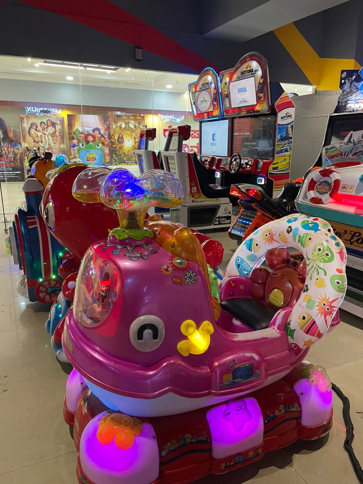 A row of colorful arcade rides for children, including a pink whale-shaped ride with cartoon sea creatures.