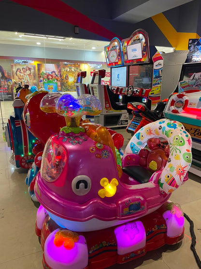 A row of colorful arcade rides for children, including a pink whale-shaped ride with cartoon sea creatures.
