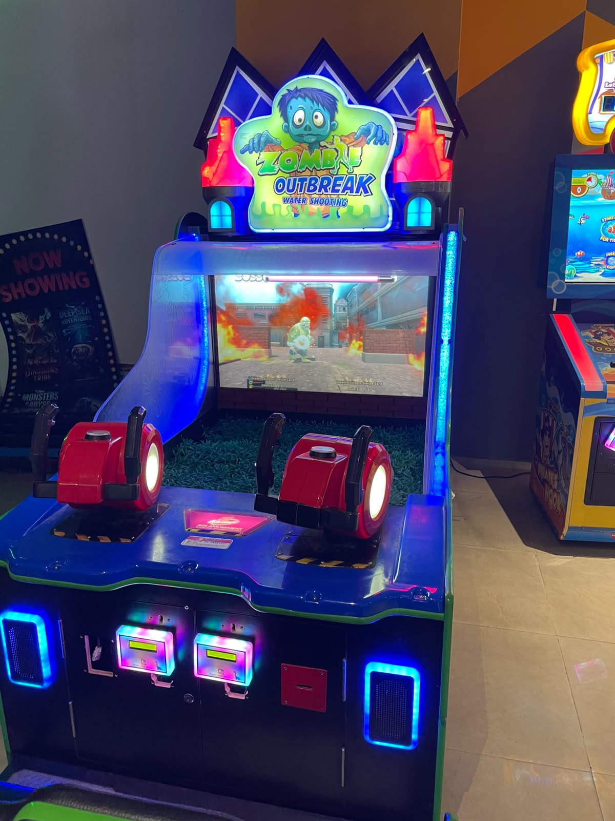 A "Zombie Outbreak" water shooting arcade game with two players aiming water guns at a screen showing a zombie in a burning city.
