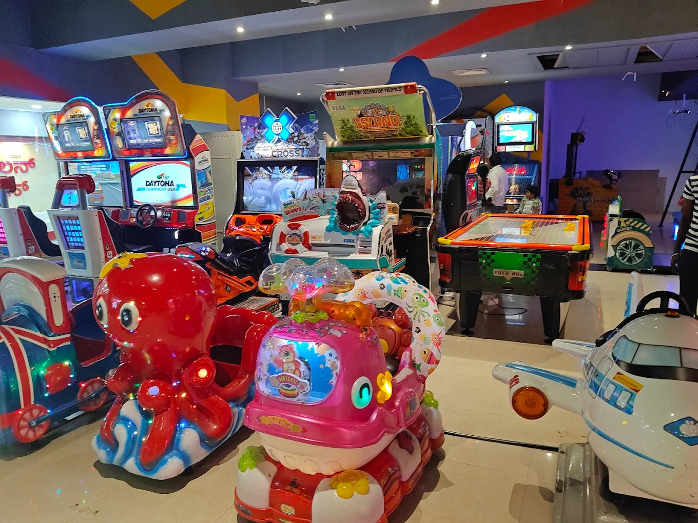 An arcade with various game machines, including racing games, a shark-themed ride, and an air hockey table. Children are visible playing.