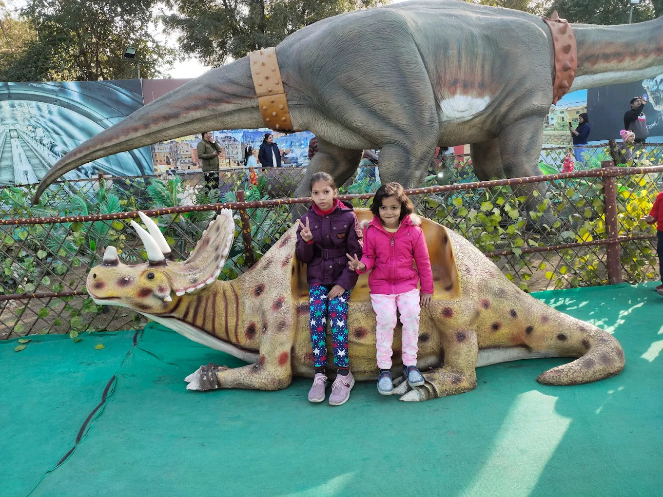 Two young girls pose on a dinosaur sculpture in front of a larger dinosaur model.