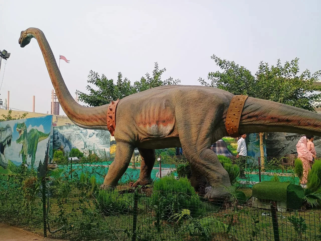A large dinosaur statue with a long neck stands in a park, with people in the background.