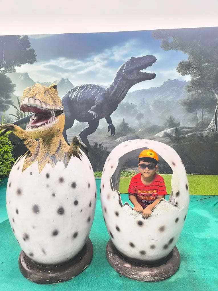 A young boy sits in a dinosaur egg prop in front of a dinosaur mural.