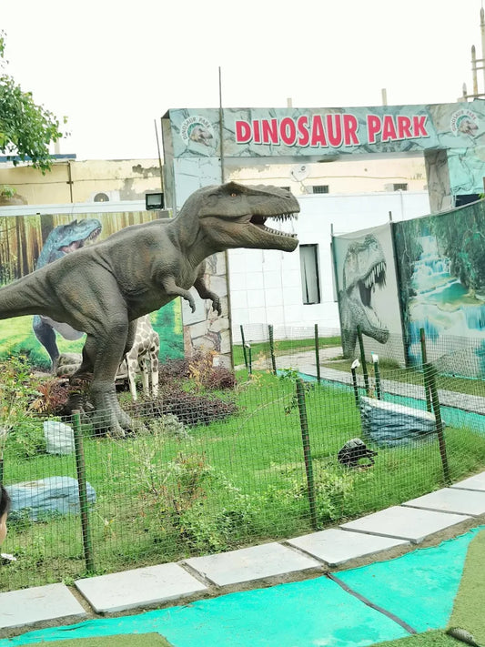 A large Tyrannosaurus Rex statue stands in front of a "Dinosaur Park" sign, with a giraffe statue in the background.