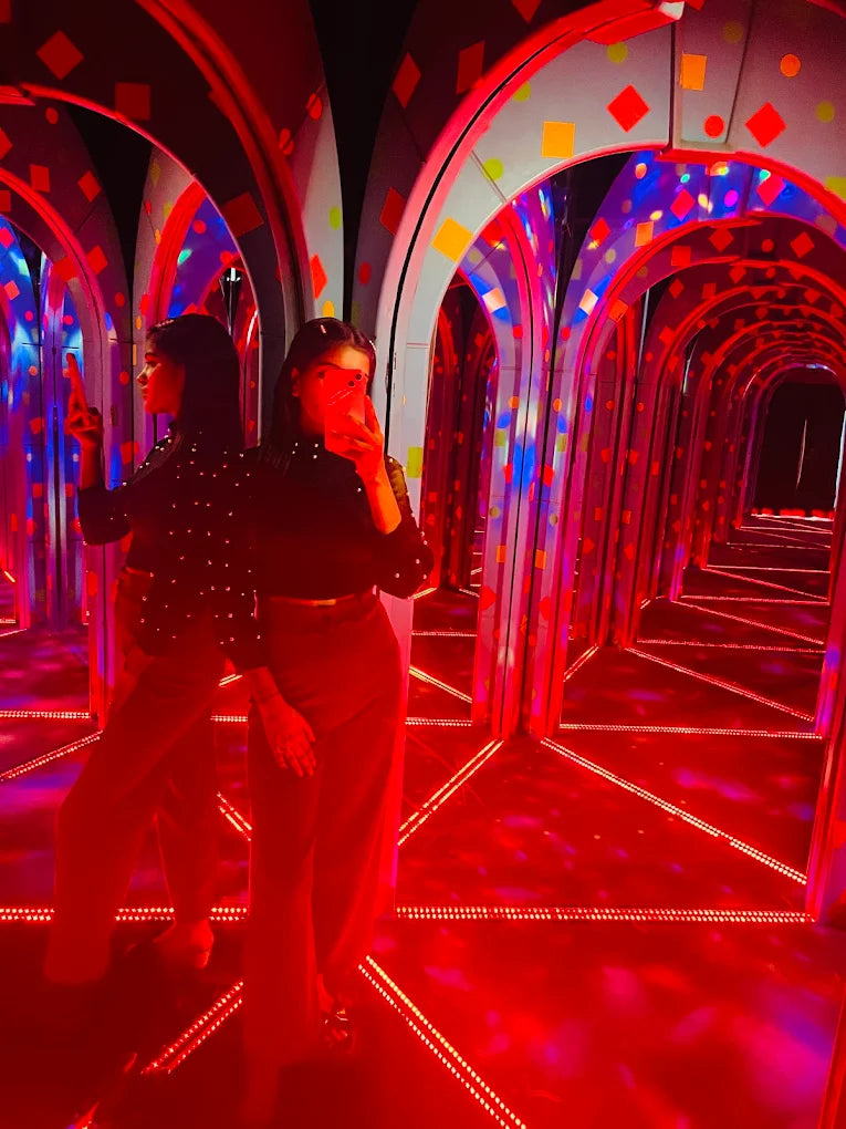 A young woman takes a selfie in a mirrored hallway with red lights and colorful arches.