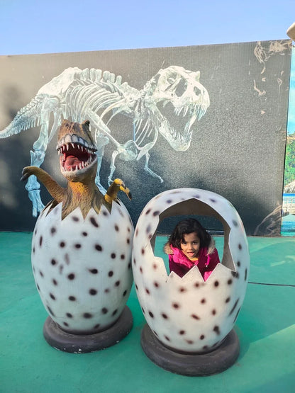 A young child peeks out of a dinosaur egg sculpture, with another egg and a dinosaur skeleton backdrop.