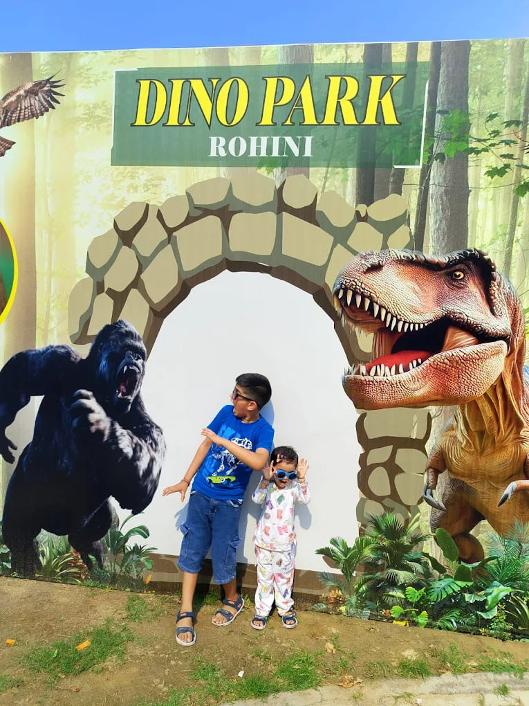 Two children pose in front of a "Dino Park Rohini" mural featuring a gorilla and a Tyrannosaurus Rex.