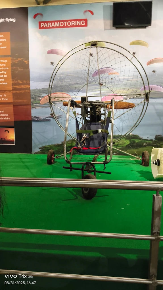 A paramotor is displayed indoors with a backdrop of paragliders in the sky.