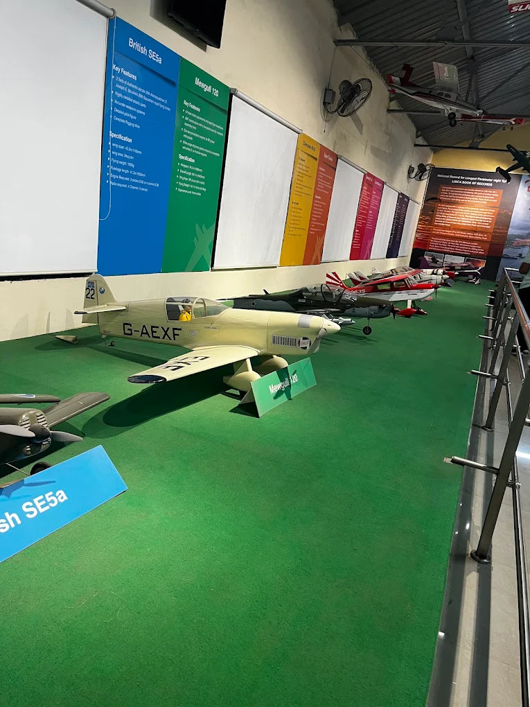 Models of vintage aircraft are displayed on a green carpet in a museum, with informational posters on the wall.