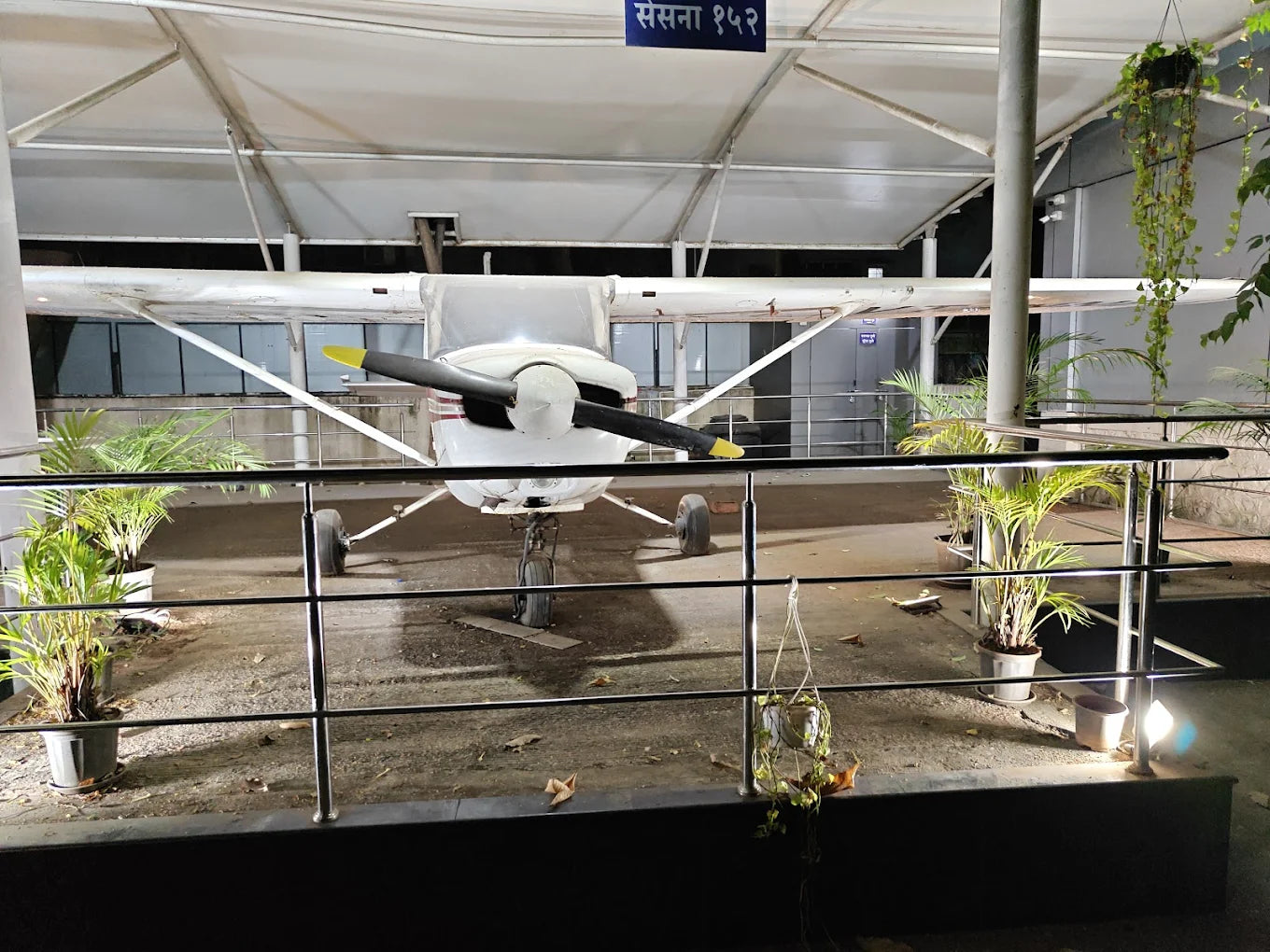 A small white airplane with a propeller is displayed indoors, surrounded by plants and a railing.