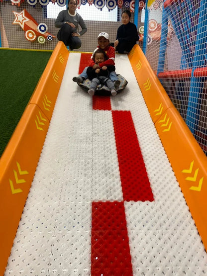 Two young children slide down a colorful indoor playground slide, with adults watching from above.