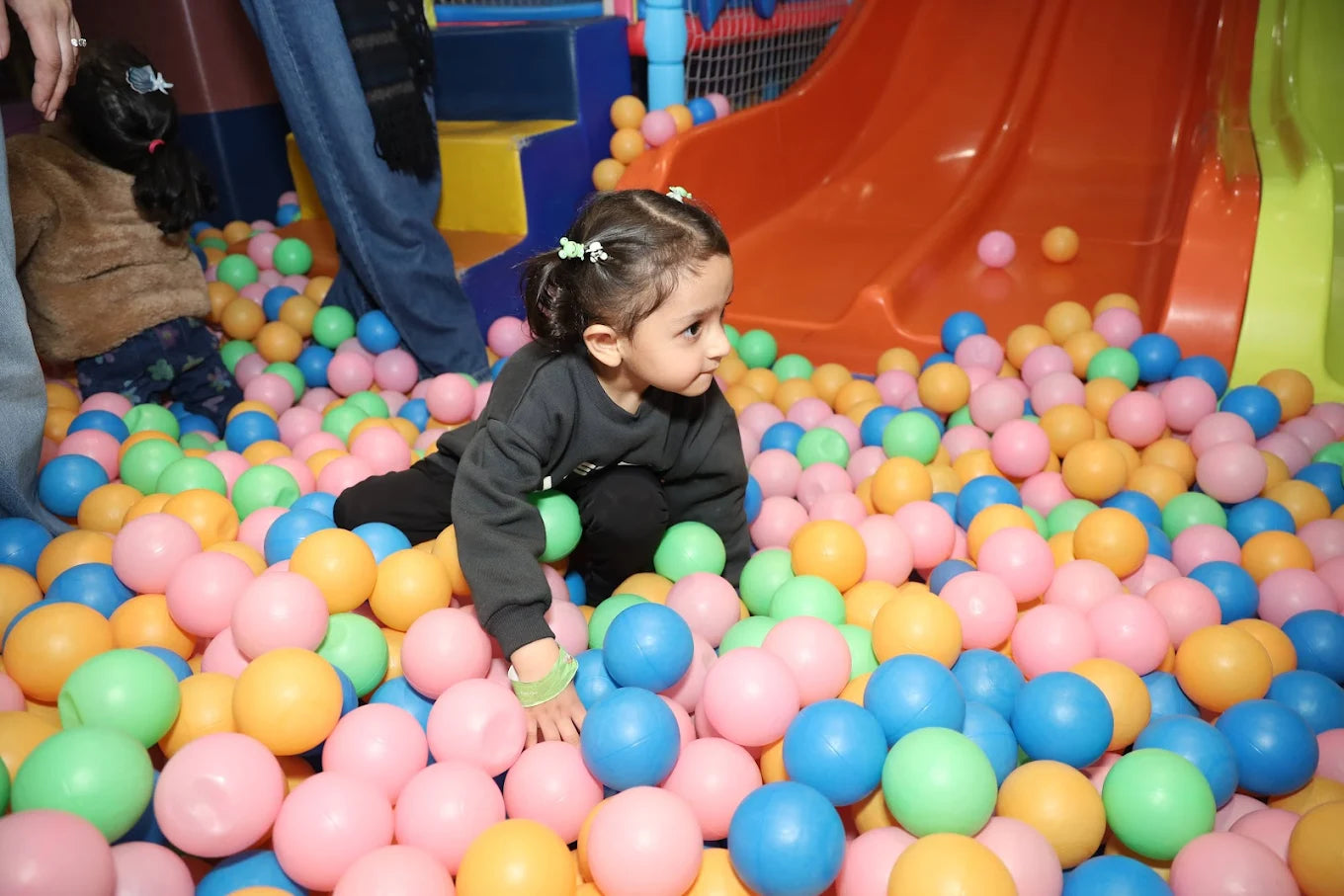 A young girl plays in a ball pit with an orange slide in the background.