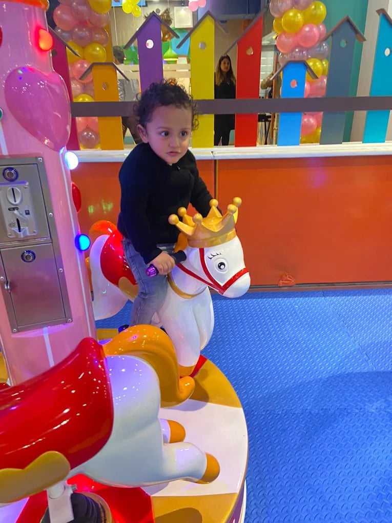 A young child rides a carousel horse in a brightly colored indoor play area.