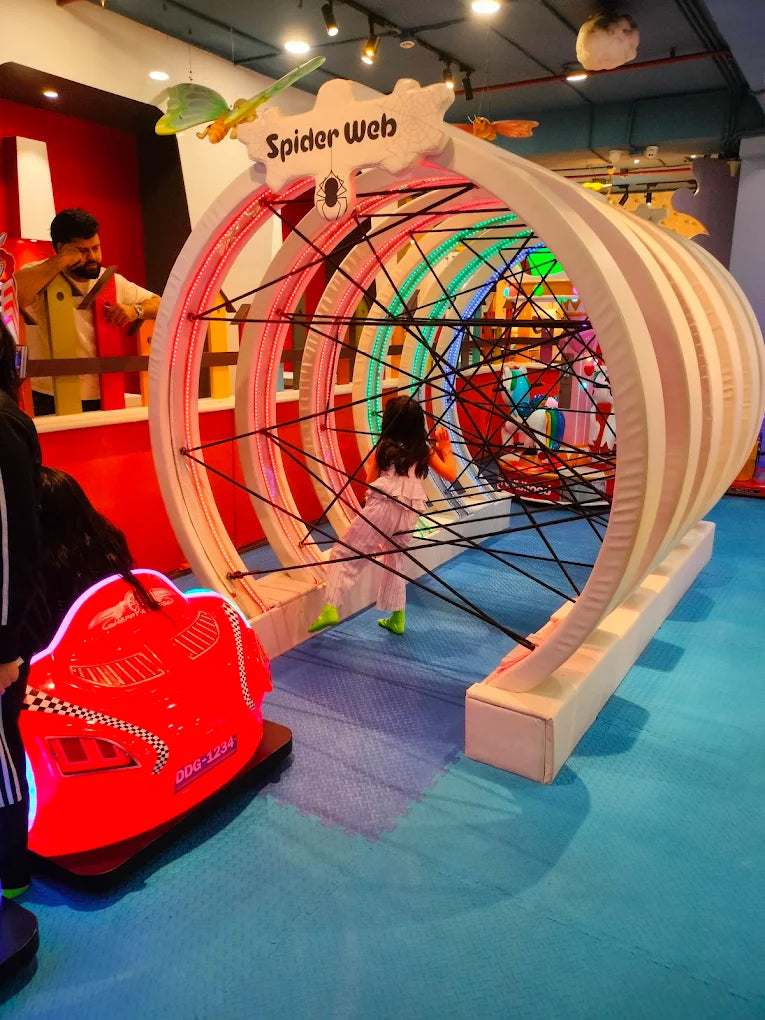 A child plays inside a large, colorful spider web structure at an indoor playground.