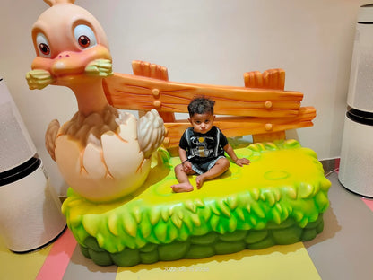 A toddler sits on a bright yellow and green grassy bench next to a large cartoon duck hatching from an egg.