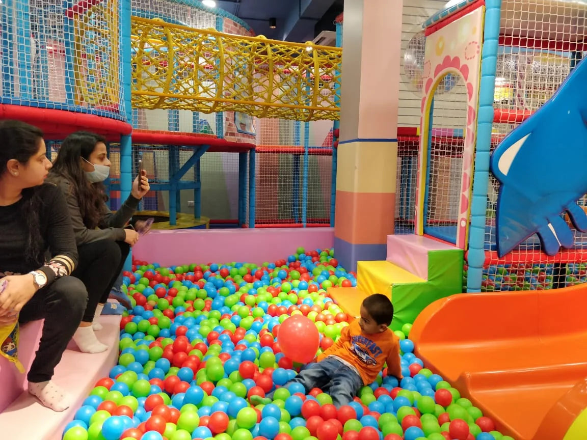 A young child plays in a ball pit at an indoor playground while two women watch.
