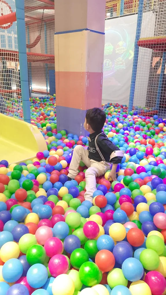 A young boy sits in a ball pit filled with colorful balls. He is looking towards a projection screen in the background.