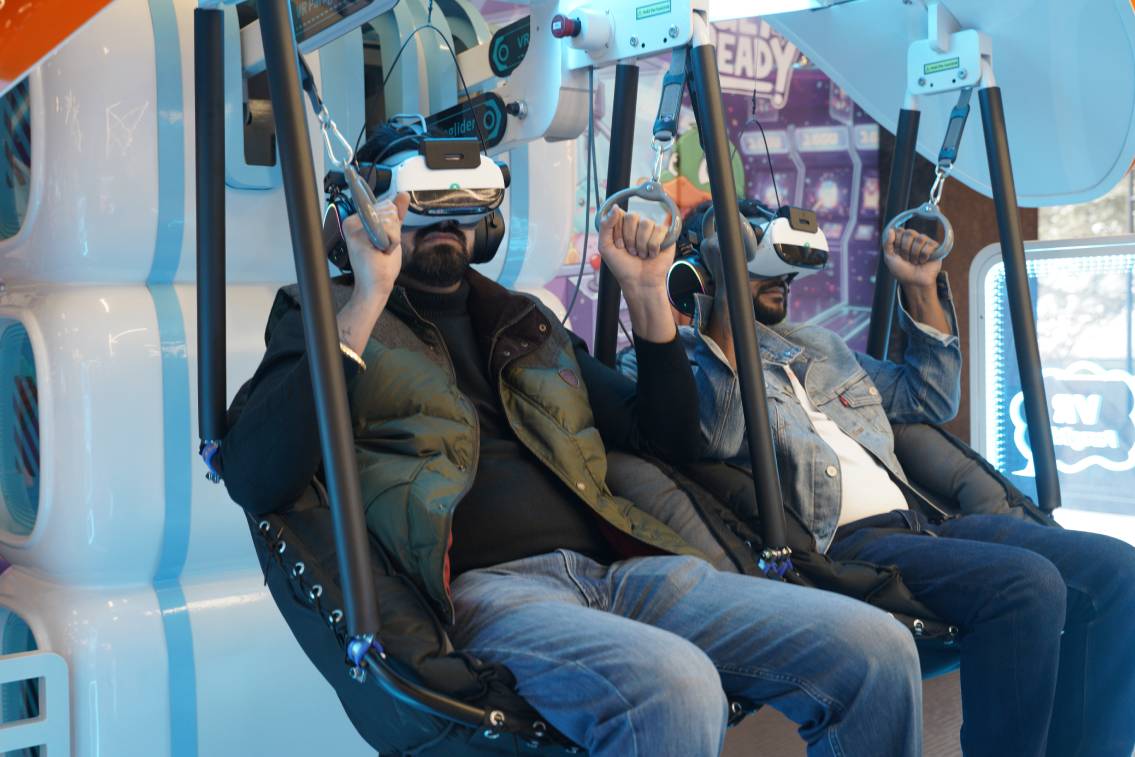 Two adults in VR headsets sit in a simulator, holding onto handles. The setting appears to be an arcade or entertainment venue.