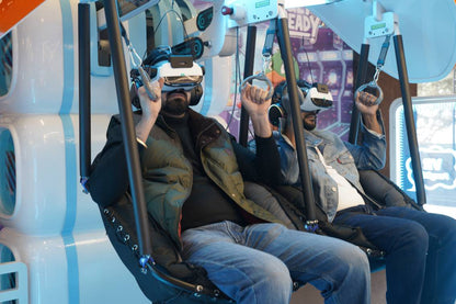 Two adults in VR headsets sit in a simulator, holding onto handles. The setting appears to be an arcade or entertainment venue.