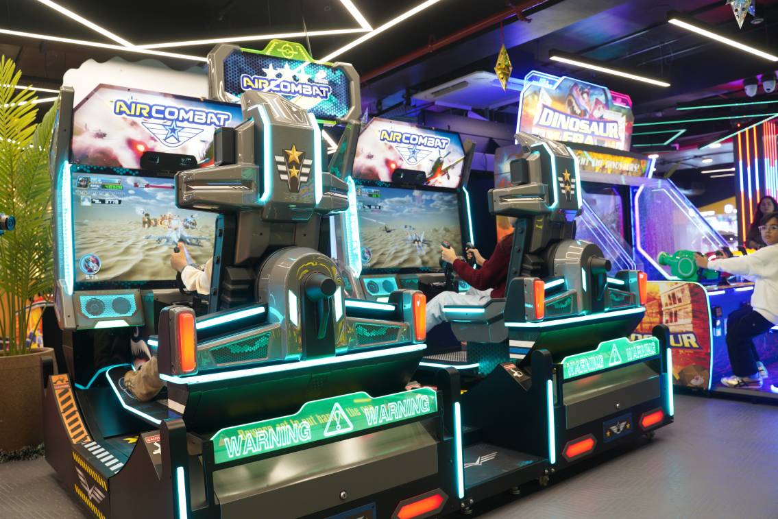 Two young people play a futuristic arcade game in a brightly lit entertainment center.