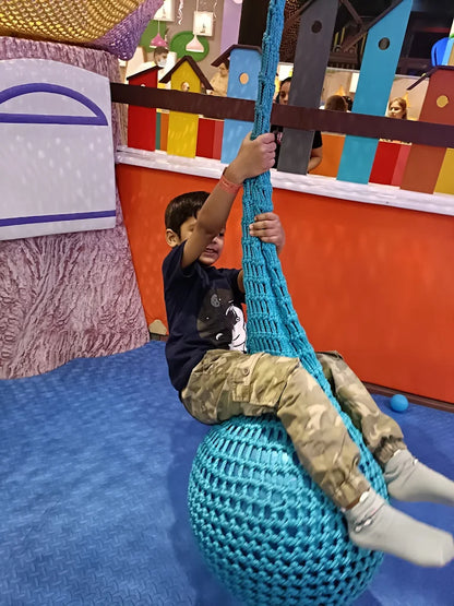 A young boy swings on a blue net seat in a colorful indoor play area.