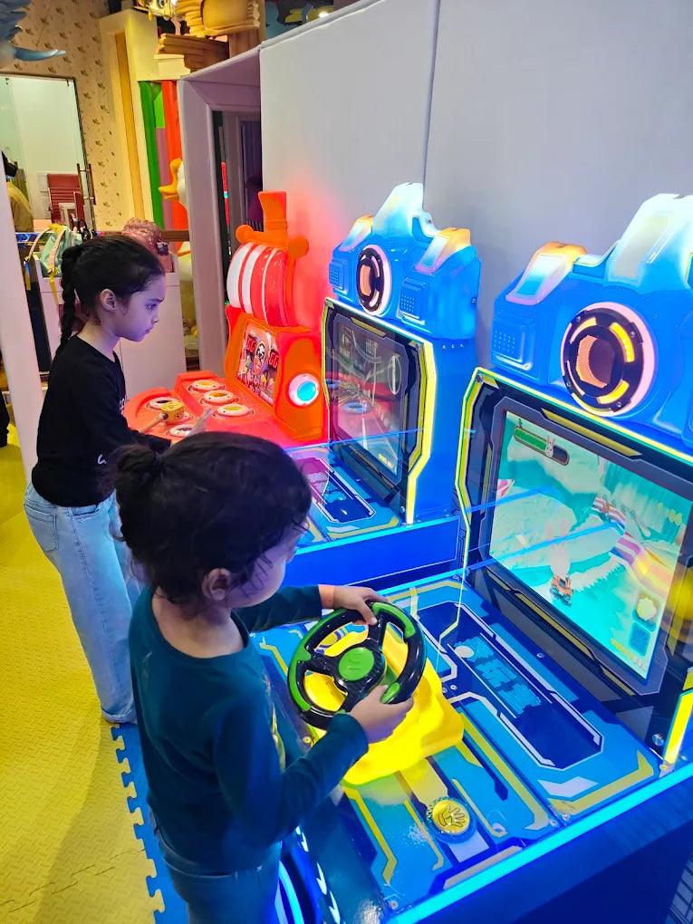 Two young girls play racing arcade games in an amusement center.