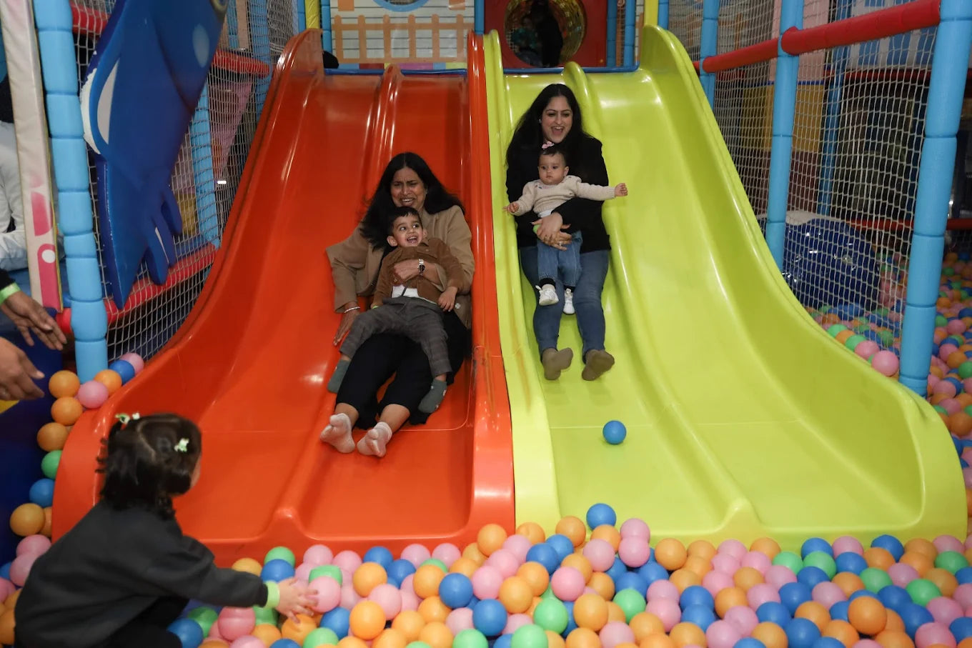 Two women and their young children slide down colorful playground slides into a ball pit.