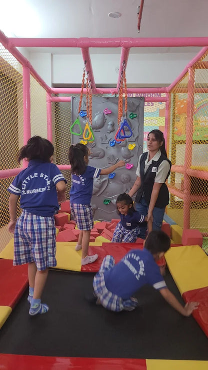 Children play in a colorful indoor playground with a climbing wall and trampoline, supervised by an adult.