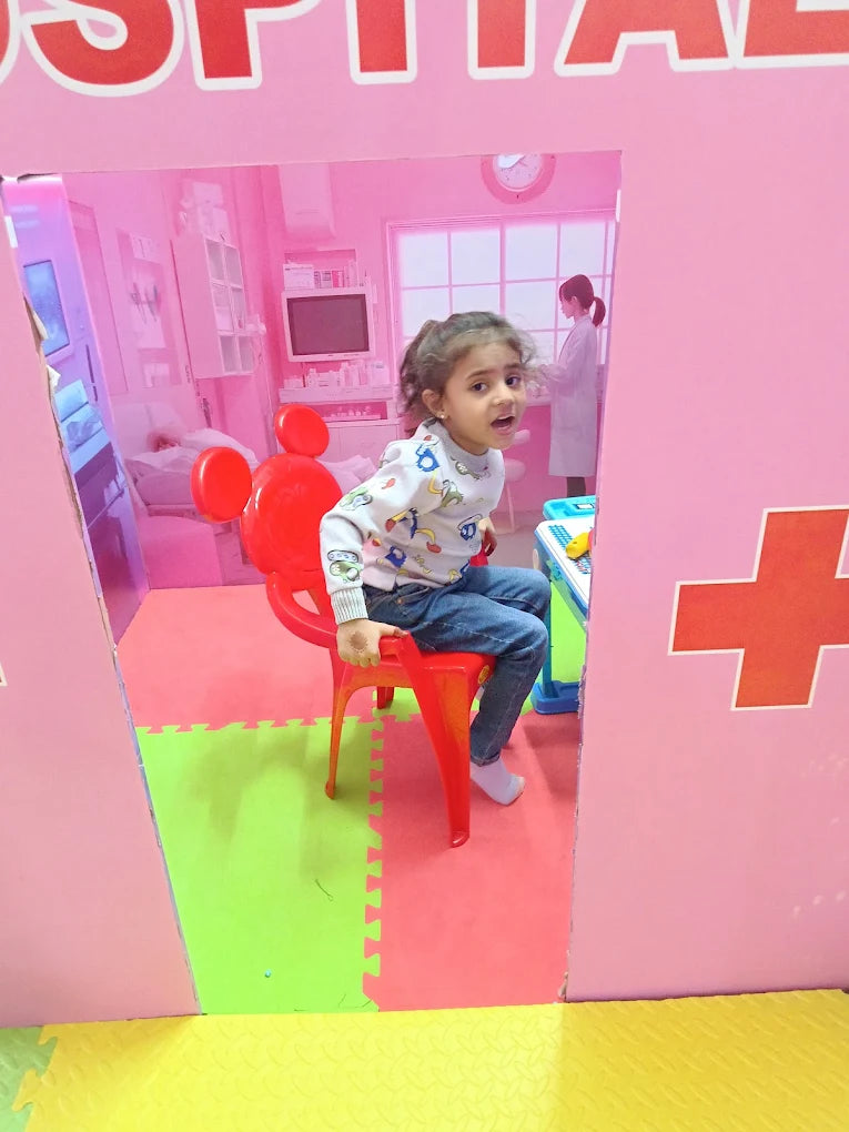 A young child sits in a toy hospital room, looking surprised. The room is pink with a red chair and a medical cross.