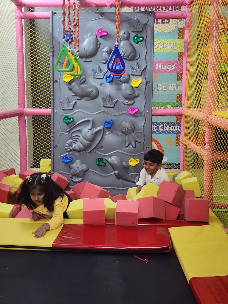 Two young children play in a ball pit in a colorful playroom with a climbing wall.