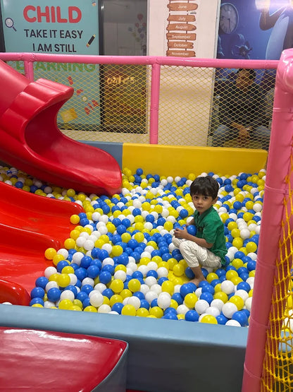 A young boy plays in a ball pit with blue, yellow, and white balls. He is sitting down, holding a blue ball.