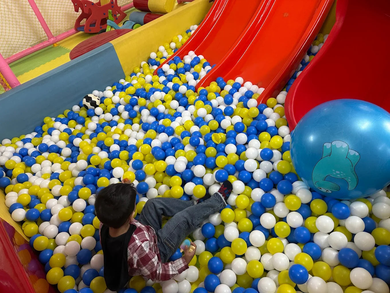 A young child plays in a ball pit with red slides and a large blue ball.