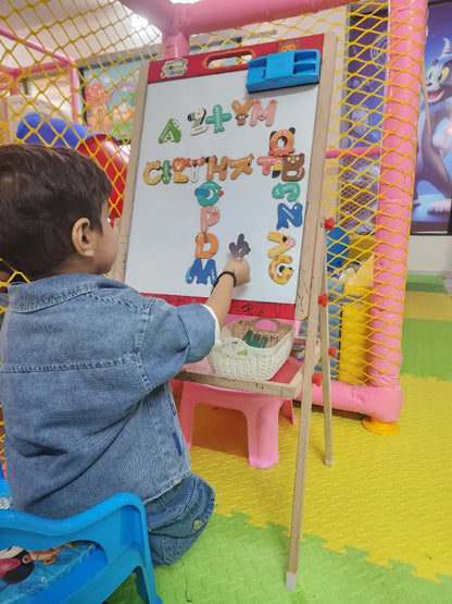 A young child in a denim outfit plays with magnetic letters on a whiteboard in a colorful indoor play area.