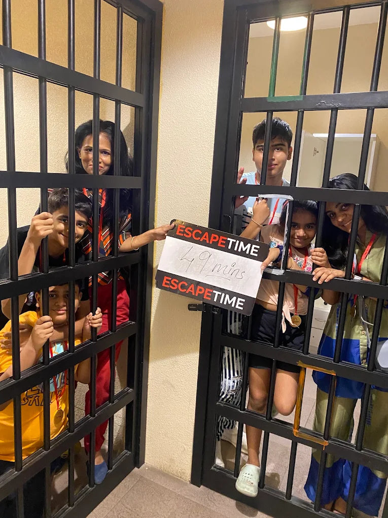 Children and adults pose behind bars at an escape room, holding a sign that says "Escape Time 49 mins."